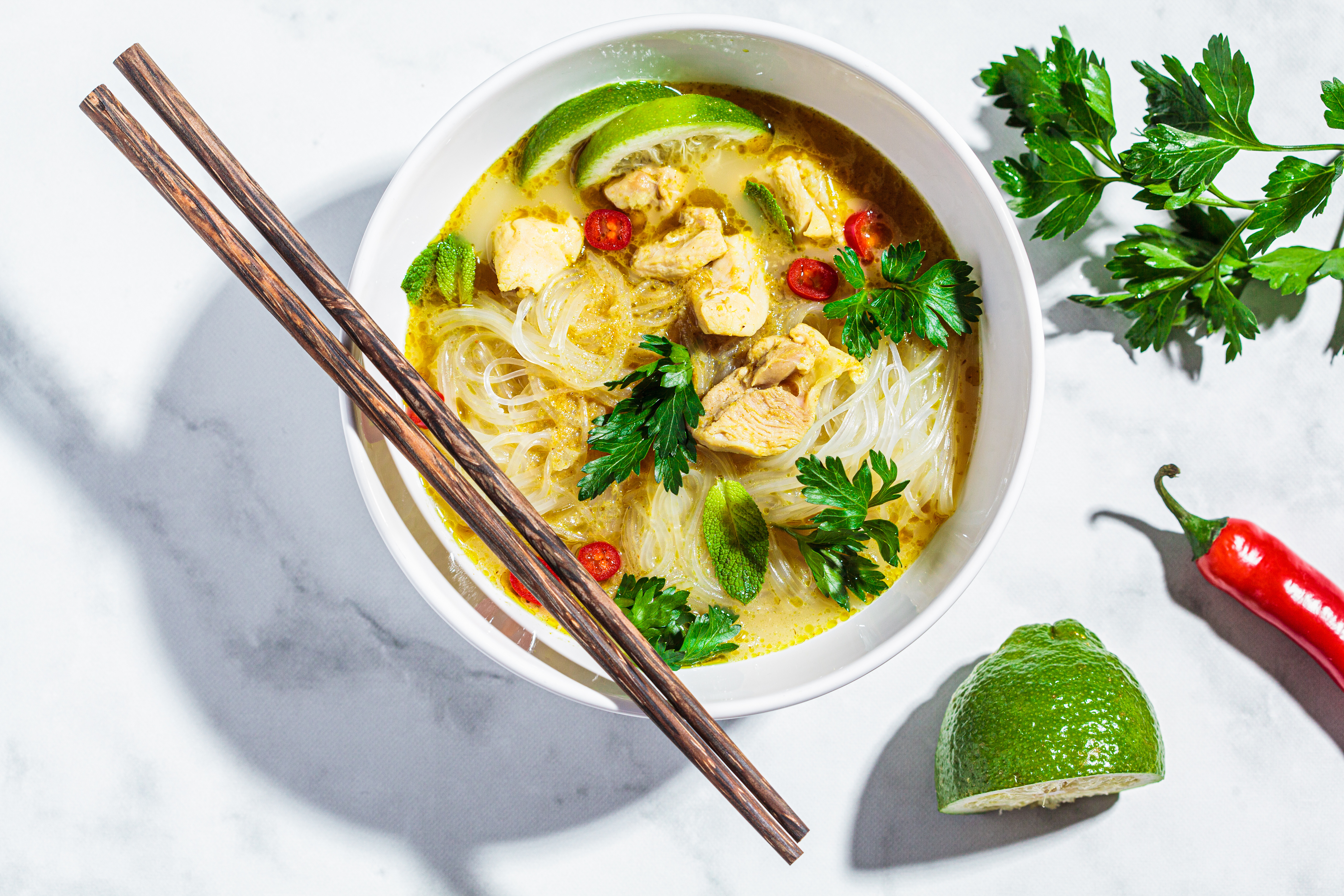 Thai green curry chicken soup with rice noodles, fresh chili, and aromatic herbs, served in a clean white bowl; a comforting and vibrant one-pot meal
